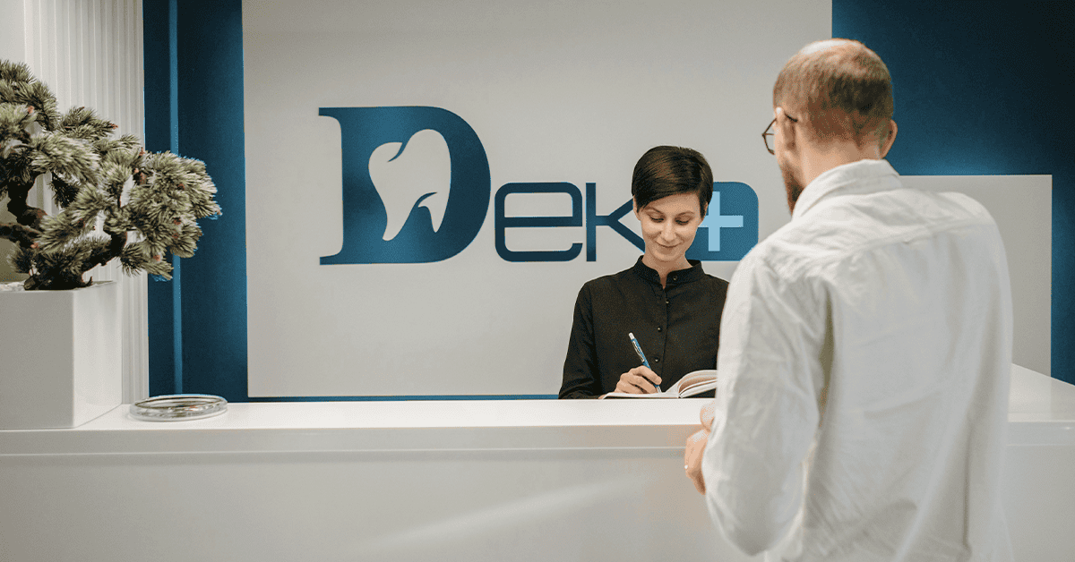 A person checking in at a dental practice reception desk, where the receptionist is writing in a book as part of the office's rebranding effort to attract more patients. OCIDM,io Branding and Digital marketing Hamilton, Toronto, Oakville, Mississauga