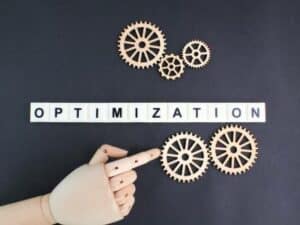 Wooden gears and a hand point to the word "OPTIMIZATION" on a tiled surface against a black background, symbolizing how web design in Toronto can transform your business online. OCIDM,io Branding and Digital marketing Hamilton, Toronto, Oakville, Mississauga