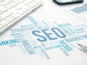 A word cloud centered around "SEO" with related terms like "marketing," "web design," and "analytics" sits next to a keyboard, pen, and smartphone on a desk, reflecting the bustling business online scene in Toronto. OCIDM,io Branding and Digital marketing Hamilton, Toronto, Oakville, Mississauga