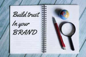 Open notebook with "Build trust in your powerhouse brand" written on the page, beside a globe, red pen, and magnifying glass on a blue wooden surface. OCIDM,io Branding and Digital marketing Hamilton, Toronto, Oakville, Mississauga