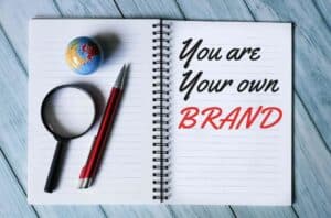 Open notebook with the text "You are Your own Powerhouse Brand," a small globe, a magnifying glass, and a red pen on a blue wooden surface. OCIDM,io Branding and Digital marketing Hamilton, Toronto, Oakville, Mississauga